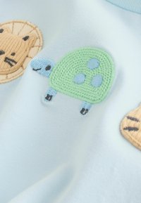 Tissu bleu clair avec motifs animaliers brodés : un lion marron, une tortue verte et une tortue bleue avec des accents beige et bleu.