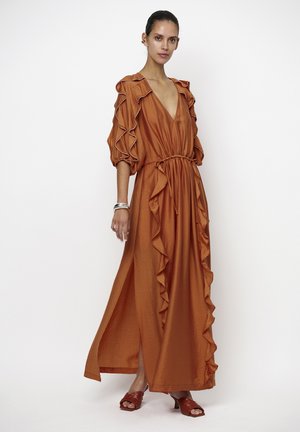 Vrouw draagt een roest-oranje maxi-jurk met ruches, diepe V-hals, pofmouwen en bijpassende open-teen sandaal met hak, tegen een effen achtergrond.