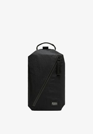Petite pochette rectangulaire noire avec fermeture éclair diagonale, poignée en tissu en haut, et petite étiquette de marque rectangulaire près du bas.