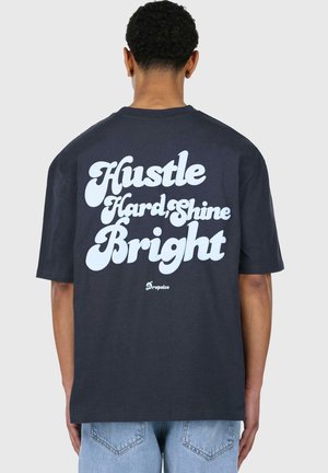 Camiseta de algodón azul marino oscuro con mangas cortas, que presenta un gran texto blanco en la parte posterior: "Trabaja Duro, Brilla Brillante", y un pequeño logotipo debajo.