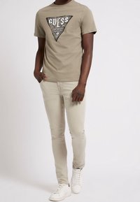 Hombre vistiendo una camiseta beige de Guess, pantalones slim-fit de color beige claro y zapatillas blancas, de pie con una mano en el bolsillo contra un fondo blanco.