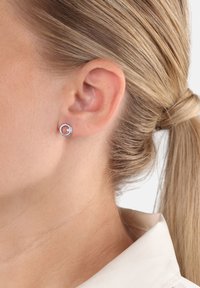 Kvinna med blont hår uppsatt som bär en liten silverf ärgad cirkulär stud earring med en liten glittrande ädelsten i vänster öra.