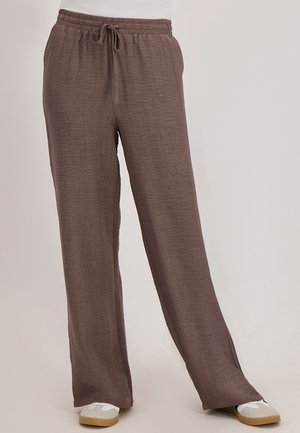 Een persoon draagt losse, getextureerde bruine broeken met een elastische tailleband en een trekkoord, gecombineerd met witte en beige sneakers op een effen achtergrond.