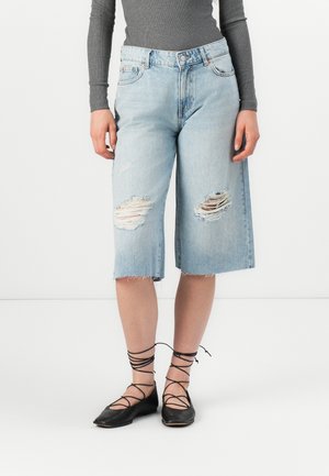 Des culottes en denim clair avec des détails déchirés et des ourlets effilochés. Associées à des ballerines noires à lacets.