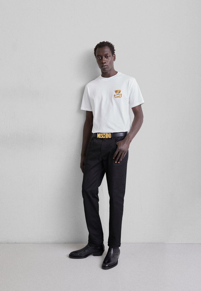 Camiseta blanca de manga corta con gráfico de oso, pantalones negros y un cinturón con el logo de Moschino. Zapatos de cuero negro texturizado completan el conjunto.