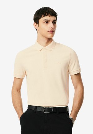 Hombre con una camisa polo beige claro de manga corta metida dentro de unos pantalones negros con cinturón negro y un reloj de pulsera plateado.