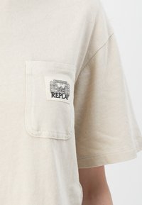 Beige T-shirt met korte mouwen van katoen, met een borstzakje waarin een zwart-wit "REPLAY" logo is afgebeeld, en een fijne, zachte textuur.