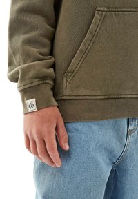 Olivgrön sweatshirt med en texturerad yta, ficka framtill, manschett med en fyrkantig logotypapplikation, bärs med ljusblå jeans.