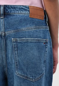 Jeans din denim albastru au o textură netedă, cusături portocalii contrastante, o etichetă din piele cu logo-ul "TOMMY JEANS" și un mic emblema tricoloră pe buzunarul din spate.