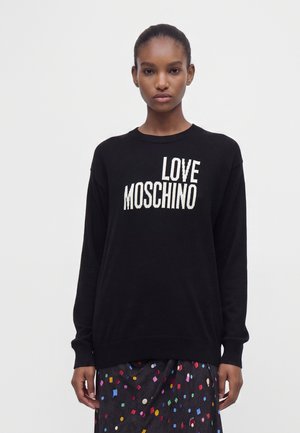 Donna che indossa un maglione nero a maniche lunghe con testo bianco "LOVE MOSCHINO" e una gonna nera con forme geometriche colorate.