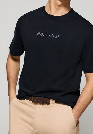 Homme portant un t-shirt noir "Polo Club" rentré dans un pantalon beige avec une ceinture marron, mains dans les poches, sur un fond uni.