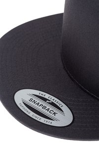 Crna snapback kapa od glatkog materijala, ravni rub, i okrugla oznaka sa detaljima "The Classics" i "Yupoong Since 1974".
