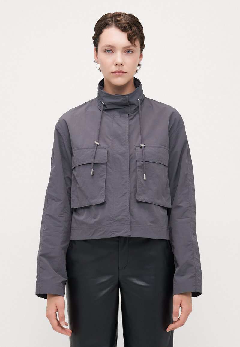 Cropped gray jacket made of lightweight fabric, featuring a high collar, two large front pockets, and adjustable drawstrings.