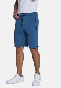 Blaue Sport Shorts mit einem Kordelzugbund, ausgestattet mit einer seitlichen Reißverschlusstasche und einem kleinen Patch-Logo am Oberschenkel. Hergestellt aus einem Baumwollmischgewebe.