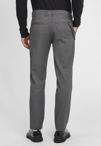 Homme portant un pantalon gris texturé avec un accessoire chaîne, une chemise noire à manches longues et des chaussures habillées noires, debout les mains dans les poches.