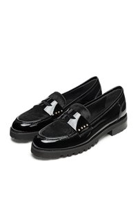 Schwarze Slipper mit einem glänzenden Lacklederkörper, einem Suede-Akzentband, metallenen Nieten und einer strukturierten Sohle für guten Halt.