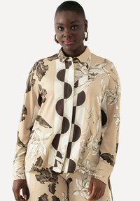 Blouse à manches longues en tissu beige avec des motifs graphiques floraux et circulaires en marron. Dotée d'un col à boutons et d'une coupe décontractée.