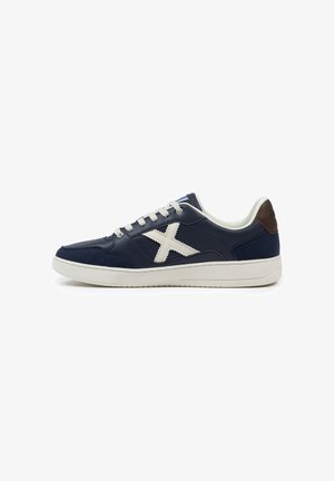 Zapatilla azul marino con suela blanca, cordones y gran logo blanco en forma de "X" en el lateral, con parche en el talón marrón y diseño bajo.