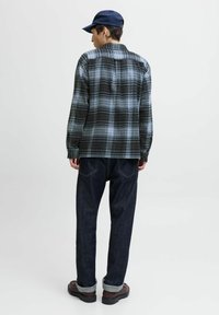 Camisa de franela a cuadros en negro y azul, vaqueros oscuros y zapatos marrones con puños remangados, vista desde atrás. Gorro azul marino colocado.