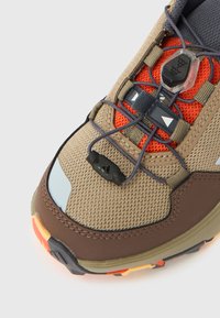 Καφέ και ταμπά αθλητικό παπούτσι με επάνω μέρος από δίχτυ, πορτοκαλί λεπτομέρειες, σύστημα κορδονιών με σφικτήρα και ανθεκτική λαστιχένια σόλα με πολύχρωμα πέλματα.