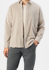 Cordhemd in hellem Beige mit Knopfleiste, langen Ärmeln und lockerem Schnitt. Kombiniert mit dunkelgrauen, schmal geschnittenen Hosen.