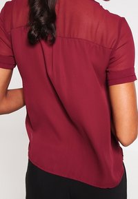 Blouse bordeaux avec des manches courtes transparentes, un ourlet arrondi et un tissu léger. Couture visible le long du dos et poignets de manches retroussés.