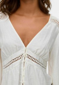 Donna che indossa una blusa bianca con scollo a V, rifinita con pizzi decorativi e bottoni sul davanti, che mostra la spalla e la parte superiore del petto.