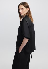 Camicia nera a maniche corte in un tessuto morbido e leggero, con taschino frontale e vestibilità rilassata. Modello in piedi con le mani in tasca davanti a uno sfondo neutro.