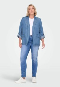 Blazer de denim azul claro con mangas enrolladas, dos bolsillos frontales y un corte relajado, combinado con jeans claros y zapatillas blancas.