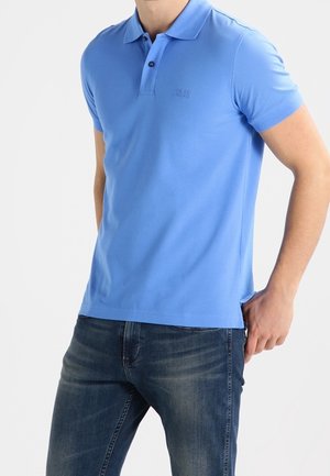 Mand iført en tætsiddende lyseblå kortærmet polo og mørkeblå jeans, stående med den ene hånd i lommen mod en hvid baggrund.