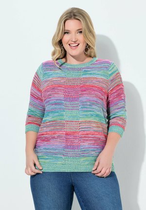 Usměvavá žena s blond vlasy nosí barevný pruhovaný pletený svetr a modré džíny, stojí před bílým pozadím.