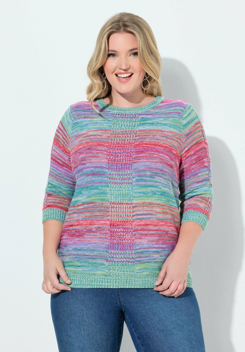 Usměvavá žena s blond vlasy nosí barevný pruhovaný pletený svetr a modré džíny, stojí před bílým pozadím.