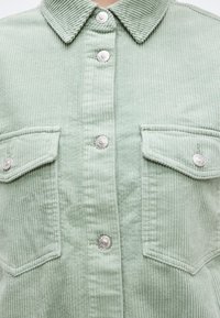 Πουκάμισο από απαλό πράσινο κορδρέ, με κάθετες ραβδώσεις, ασημένια κουμπιά κρατημένα με πάτημα στην μπροστινή πλευρά και δύο τσέπες στο στήθος με καλύμματα που κλείνουν με κουμπί.