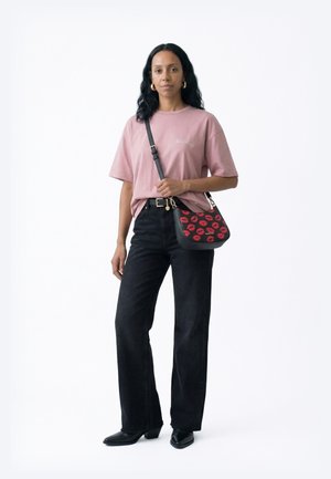 Woman with long dark hair wearing pink oversized T-shirt, black high-waist jeans, black ankle boots, gold hoop earrings, and black crossbody bag with red lips.