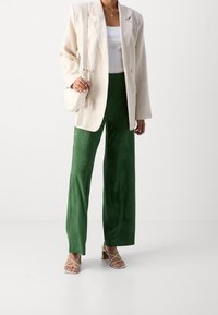 Světle béžové oversized sako, bílý žebrovaný top na ramínka, zelené kalhoty širokých nohavic s listovým vzorem a sandály na blokovém podpatku s pásky.