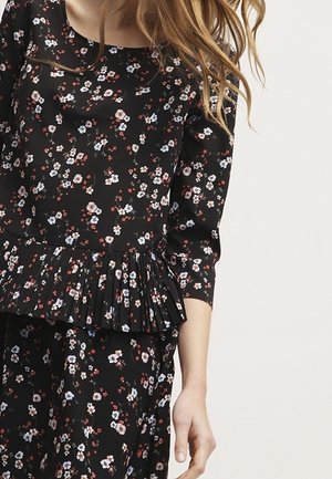 Kobieta ubrana w czarną sukienkę w kwiaty z długim rękawem i plisowanym detalem w talii, ręka swobodnie opuszczona wzdłuż boku, jasne tło.