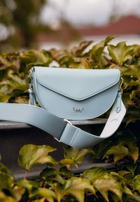 Světle modrá kožená kabelka přes rameno s klopou na zapínání, stříbrným kováním a nastavitelným popruhem. Hladká textura s minimalistickým designem.