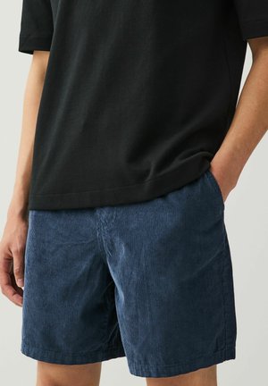Navyblå kordfløjlsshorts med struktureret stof og en afslappet pasform, kombineret med en sort T-shirt med korte ærmer. Hånd i lomme-detalje synlig.