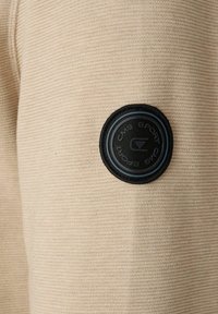 Beige gebreide stof met een geribbeld patroon, voorzien van een ronde zwarte patch met blauwe tekst met de label "CMS SPORT" aan de linker zijde.