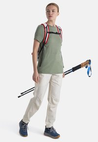 Abbigliamento da escursionismo composto da una maglietta verde chiaro, pantaloni crema con tasche e uno zaino nero con accenti rossi. Include bastoncini da trekking con impugnature in sughero.
