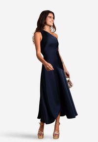 Robe en satin bleu marine avec un design à une épaule, silhouette en A et ourlet fluide. Associée à des sandales compensées beiges et un petit sac.