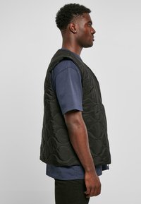 Zwarte gewatteerde vest met een hoge hals, mouwloos ontwerp en een rits aan de voorkant. G gedragen over een marineblauw shirt met korte mouwen.