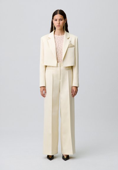 Blazer ajusté de couleur crème avec une coupe cropped, associé à un pantalon large taille haute. Haut transparent à motifs floraux en dessous. Chaussures noires à talons pointus.