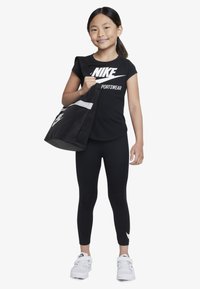 Musta Nike-t-paita, jossa valkoinen logo ja teksti, mustat legginssit, kädessä musta laukku valkoisella raidalla ja jalassa valkoiset tennarit.
