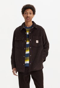 Brown corduroy jacket with two breast pockets, button-up front, paired with a multicolored striped t-shirt, worn with matching pants.