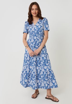 Frau trägt ein blaues geblümtes Midikleid mit kurzen Ärmeln und braunen Riemchensandalen, steht vor einem schlichten hellen Hintergrund.