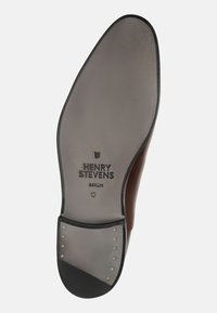 Sole di una scarpa in pelle marrone con superficie liscia, marchio impreziosito in rilievo e borchie metalliche decorative vicino al tallone. Indicato numero 10.