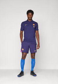 Dres FC Barcelone, ki vključuje temno vijolično polo majico z grbom in rumenimi akcenti, v kombinaciji z usklajenimi kratkimi hlačami in modrimi nogavicami.