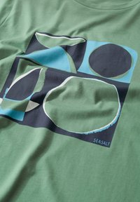 Organická bavlněná tričko v zelené barvě se stylizovanými tvary v modré a černé. Design obsahuje křivkové linie a kontrastní barvy. Viditelný štítek značky.