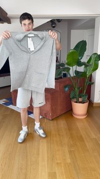 Šviesiai pilka, raukšlėta megztinė su dideliuapykakle, sagomis ir dviem priekinėmis kišenėmis, laikoma asmens neformaliame aplinkoje.